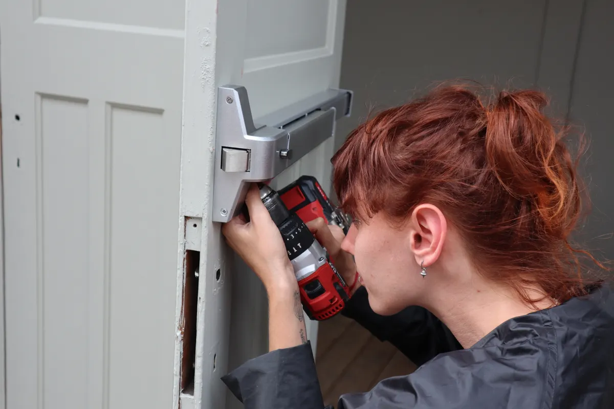 Cerrajera instalando con un taladro inalámbrico una barra antipánico en una puerta de madera blanca