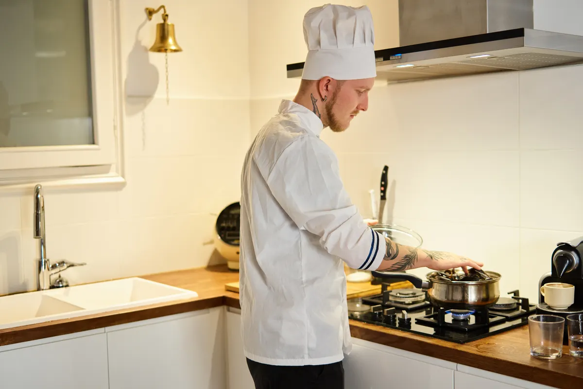 Cocinero tatuado con gorro pendiente de una olla en la placa de una cocina moderna