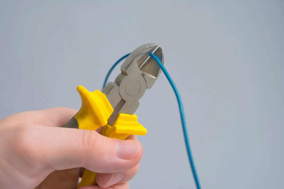 Detalle de unos alicates en una mano cortando un cable azul, con fondo gris