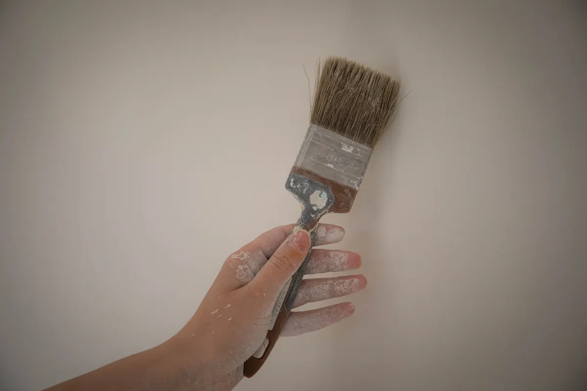 Mano sujetando una brocha con pintura blanca frente a una pared lisa