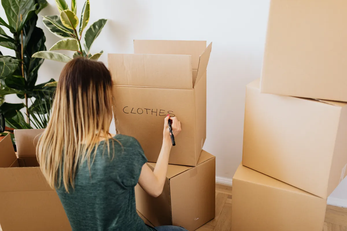 Mujer de espaldas escribiendo la palabra ropa en una caja de cartón mientras prepara la mudanza
