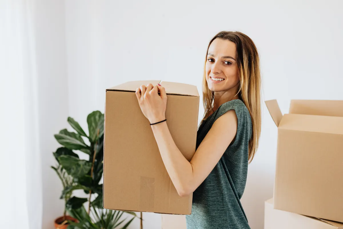 Mujer sonriente con camisa verde llevando una caja de cartón durante la mudanza a su nueva casa