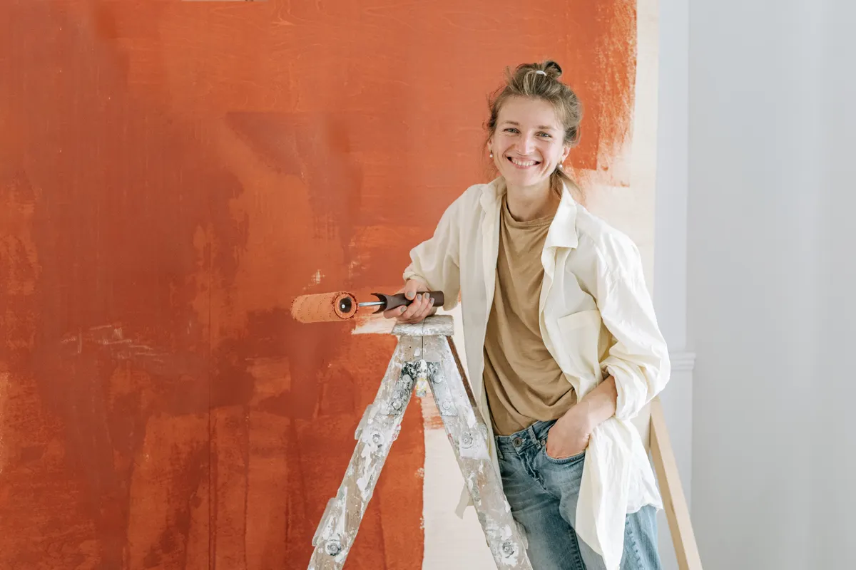 Mujer sonriente con ropa informal subida a una escalera pintando una pared naranja