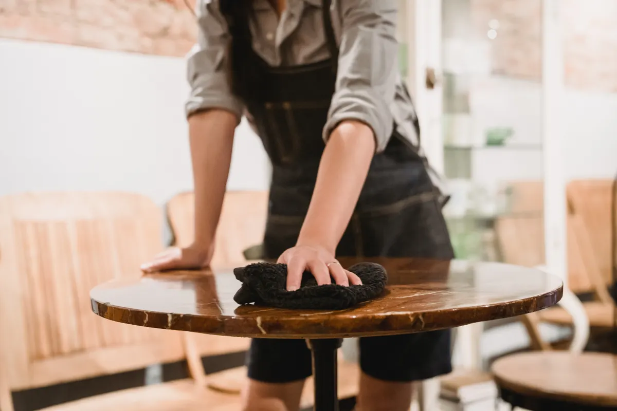 Persona con delantal limpiando una mesa de madera en una cafetería acogedora