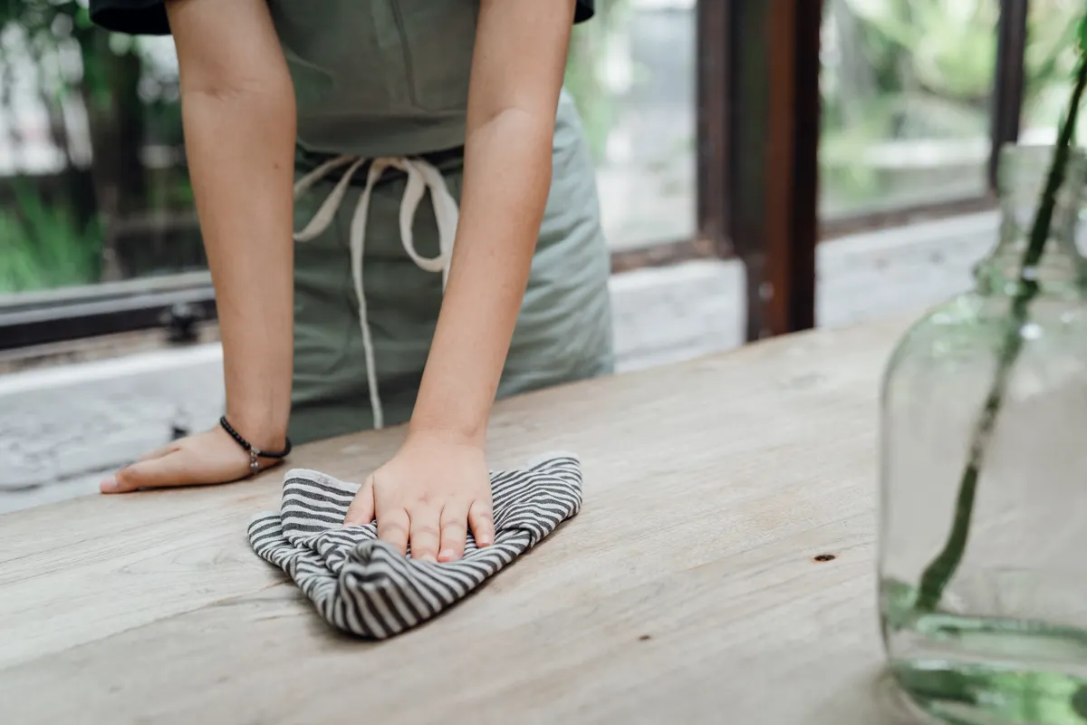 Persona con delantal verde pasando un paño de rayas por una mesa de madera junto a una ventana