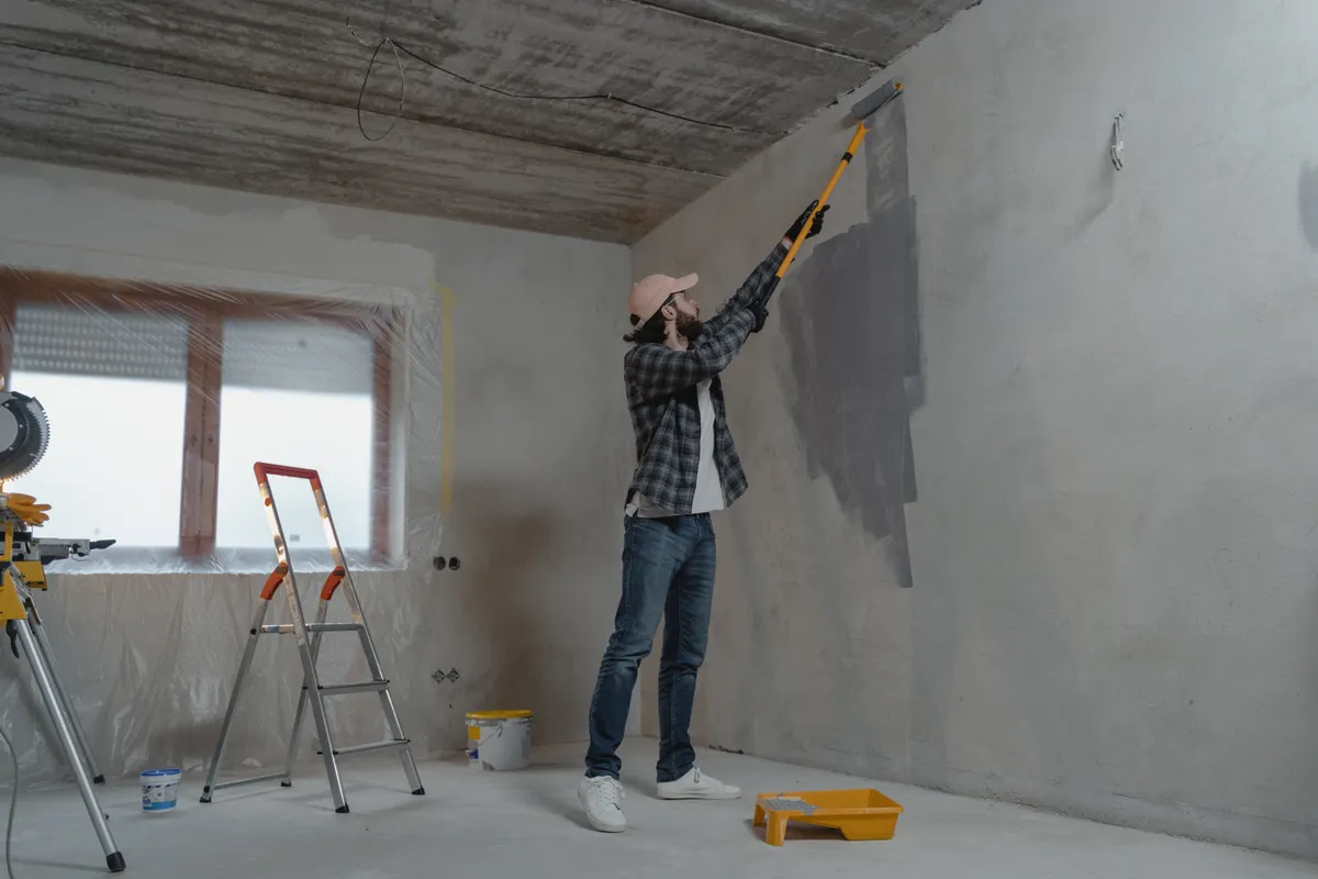 Pintor pasando el rodillo con alargador por una pared a medio pintar de gris en una vivienda en obras