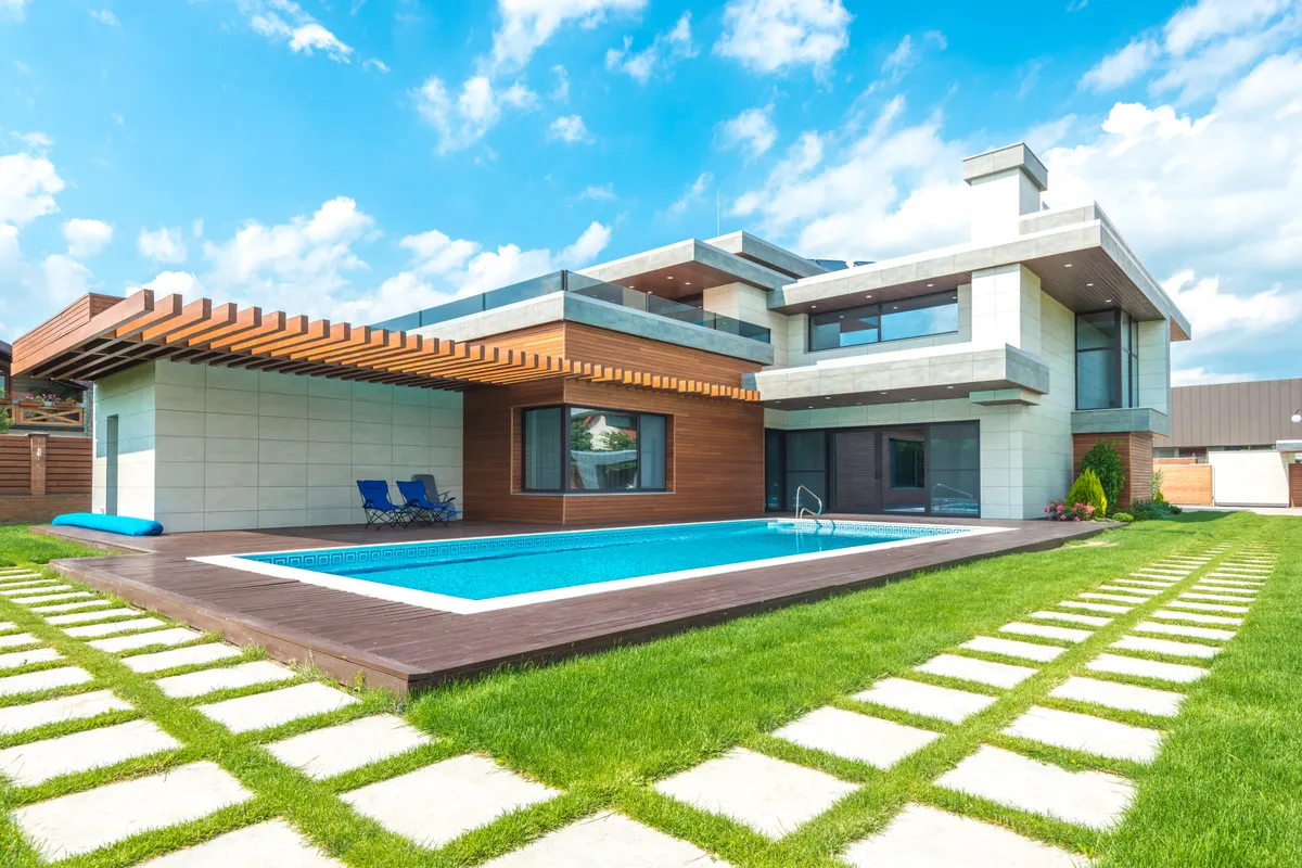Piscina de obra rectangular con tarima de madera, césped y camino de losas junto a una casa moderna
